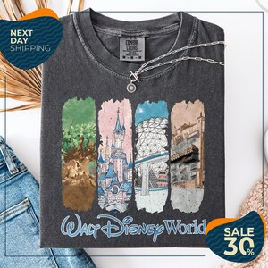 a gray t-shirt with a graphic design depicting various Disney World landmarks, including the Walt Disney World resort, the Wood Tower, and the Epcot Pavilion. The t-shirt is displayed on a white background, and there are jeans and a necklace visible in the foreground.