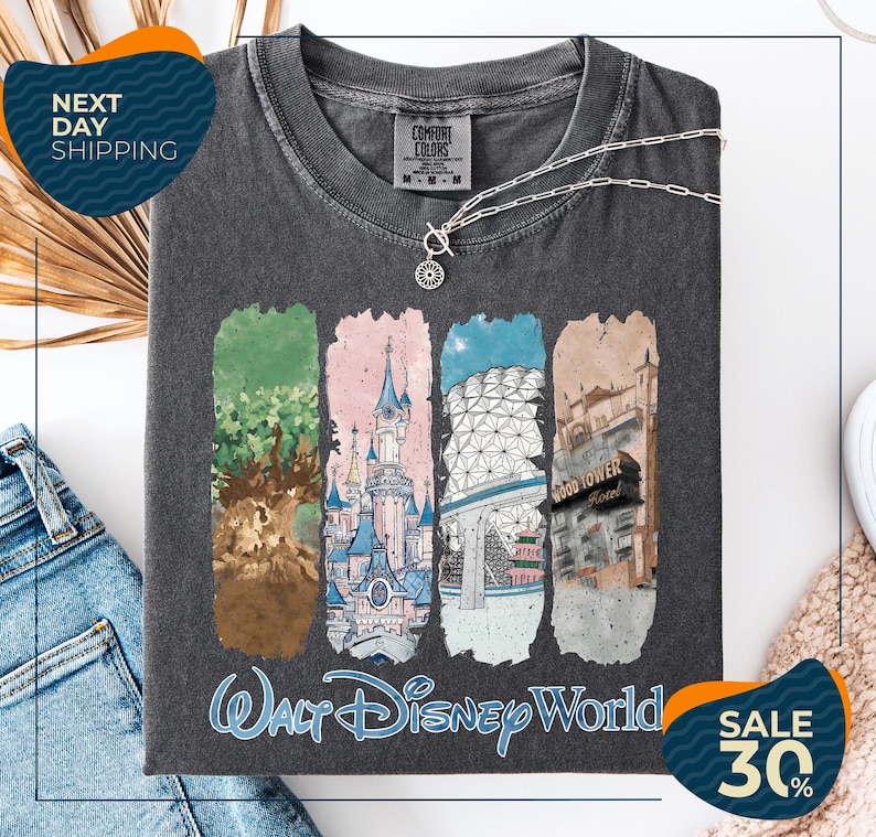 a gray t-shirt with a graphic design depicting various Disney World landmarks, including the Walt Disney World resort, the Wood Tower, and the Epcot Pavilion. The t-shirt is displayed on a white background, and there are jeans and a necklace visible in the foreground.