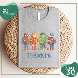 A gray t-shirt with a graphic design featuring four cartoon characters, including Spider-Man, Iron Man, Hulk, and Captain America, along with the name Theodore printed on the shirt.