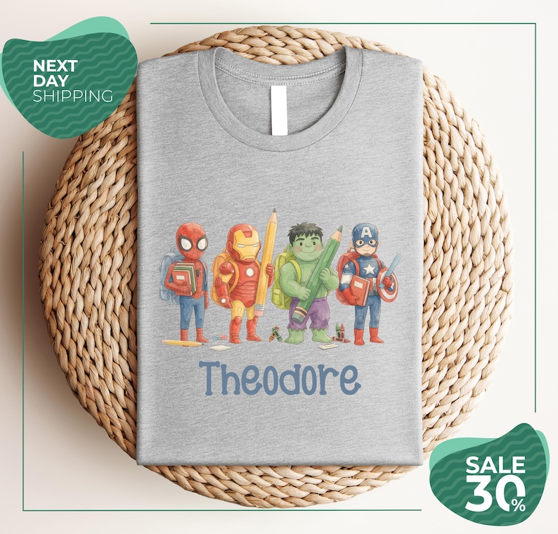 A gray t-shirt with a graphic design featuring four cartoon characters, including Spider-Man, Iron Man, Hulk, and Captain America, along with the name Theodore printed on the shirt.