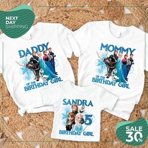 three white t-shirts with the same design, each with a different character from the Disney movie Frozen. The design includes the characters Elsa, Anna, Olaf, and other popular characters from the movie. The t-shirts are arranged on a bed of straw, and there is a sale sign indicating a 30% discount on the t-shirts.