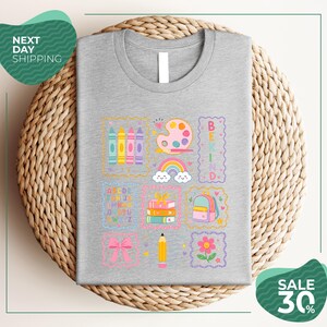 a gray t-shirt with a colorful and whimsical design, including various school supplies and educational elements such as crayons, pencils, books, and a backpack. The t-shirt is displayed on a woven basket, and there are two green circular signs in the image, one on the left side and one on the right side, which appear to be related to shipping or a sale.