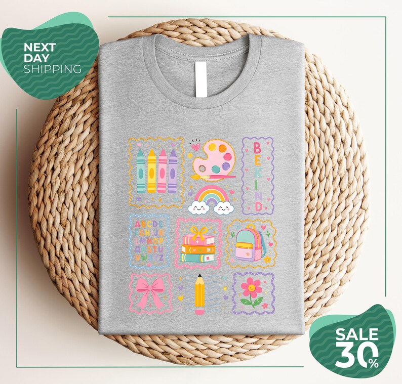 a gray t-shirt with a colorful and whimsical design, including various school supplies and educational elements such as crayons, pencils, books, and a backpack. The t-shirt is displayed on a woven basket, and there are two green circular signs in the image, one on the left side and one on the right side, which appear to be related to shipping or a sale.