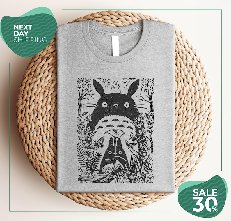 a gray t-shirt with a black and white graphic design depicting a cat surrounded by various plants and flowers. The t-shirt is placed on a woven placemat, and there are two green circular sale signs in the image.