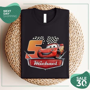 A black t-shirt with a graphic design featuring a red car and checkered flags, along with the text "Michael" and the number "5". The t-shirt is displayed on a woven placemat, and there are promotional texts for shipping and a sale offer.