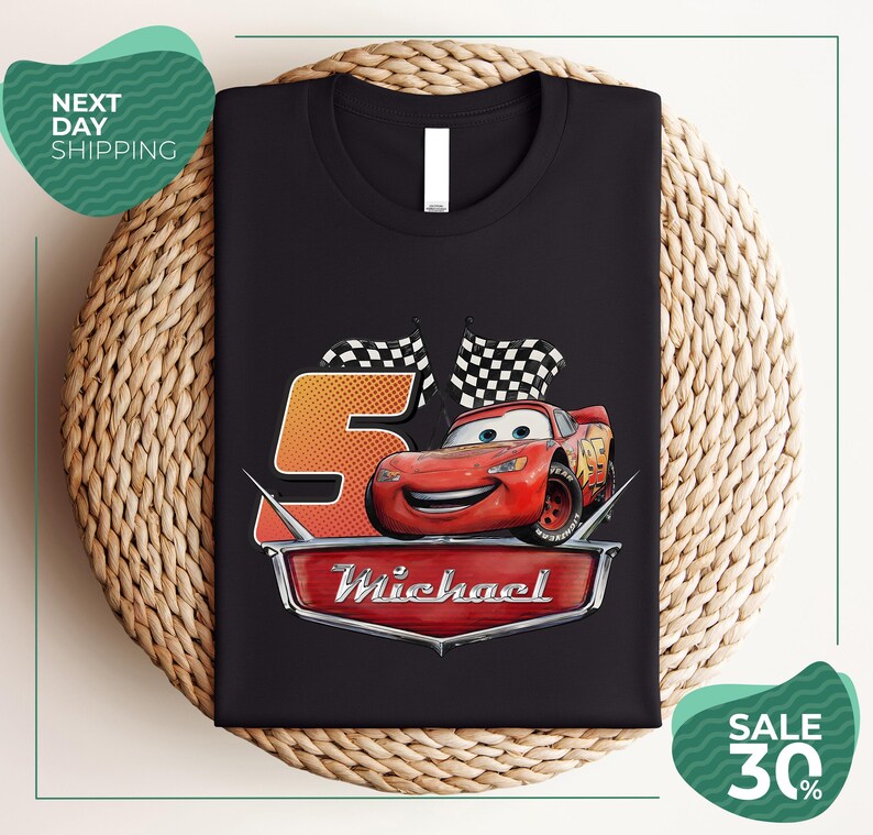 A black t-shirt with a graphic design featuring a red car and checkered flags, along with the text "Michael" and the number "5". The t-shirt is displayed on a woven placemat, and there are promotional texts for shipping and a sale offer.