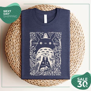 a blue t-shirt with a white graphic design depicting a cat surrounded by various natural elements, such as leaves, flowers, and mushrooms. The t-shirt is placed on a woven placemat, and there are two green circular signs in the image. One sign advertises "Next Day Shipping" and the other sign advertises a "SALE 30%".