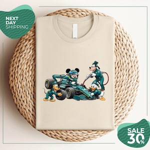 May include: A beige t-shirt featuring a graphic of Mickey Mouse in a racing helmet sitting in a green Formula 1 car, with Goofy, Pluto, and Donald Duck dressed in matching green pit crew uniforms performing maintenance on the vehicle.