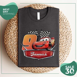 A gray t-shirt with a graphic design featuring a red car and checkered flags, along with the text "James" and the number "9". The t-shirt is displayed on a woven placemat, and there are two green sale signs in the image.