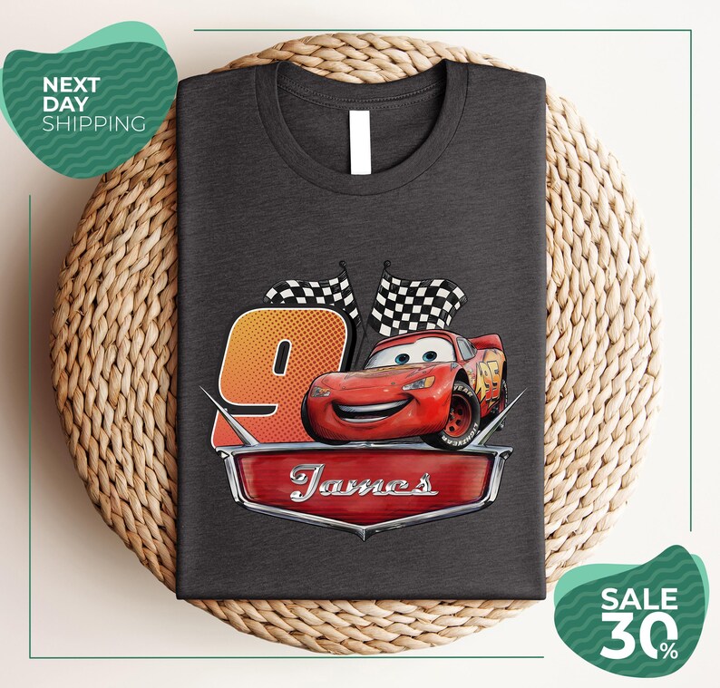 A gray t-shirt with a graphic design featuring a red car and checkered flags, along with the text "James" and the number "9". The t-shirt is displayed on a woven placemat, and there are two green sale signs in the image.