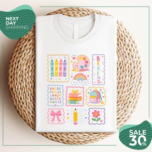 A white t-shirt with a colorful graphic design featuring various school-related items such as crayons, a backpack, a pencil, and a rainbow. The design also includes the words "BEKIND" and "SALE 30%".