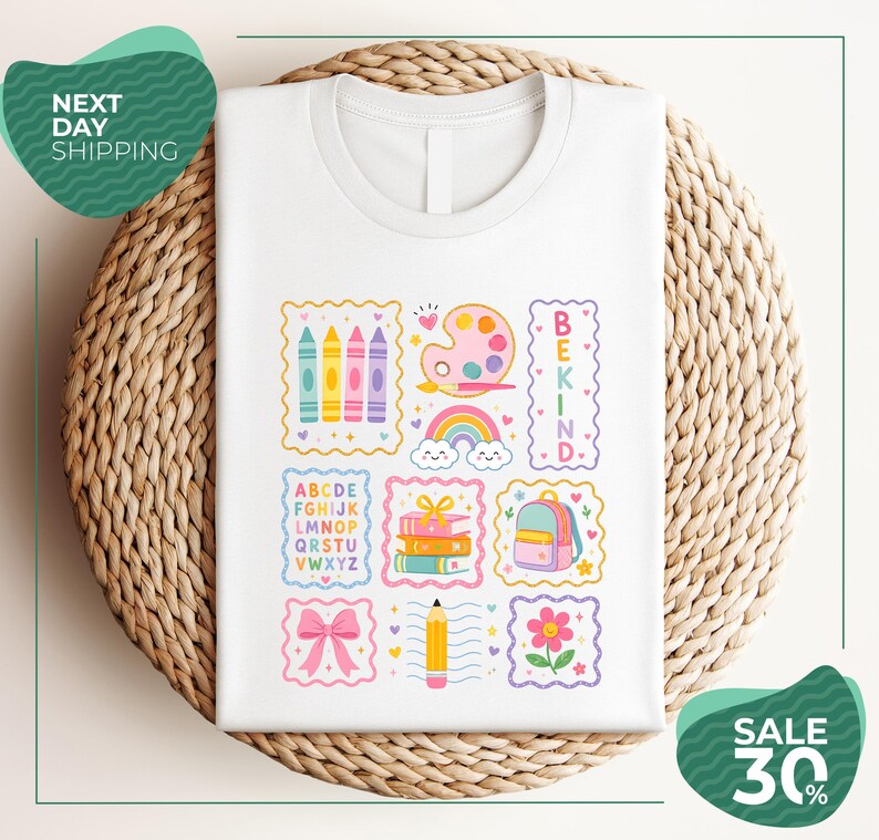 A white t-shirt with a colorful graphic design featuring various school-related items such as crayons, a backpack, a pencil, and a rainbow. The design also includes the words "BEKIND" and "SALE 30%".
