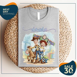 a gray t-shirt with a cartoon illustration of characters from the Toy Story franchise, including Woody, Buzz Lightyear, and an alien character. The t-shirt is displayed on a woven placemat, and there are two sale signs indicating a 30% discount on the item.