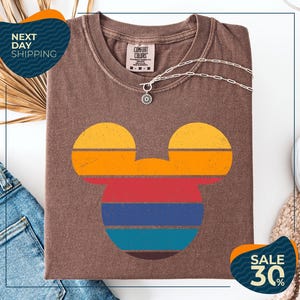a brown t-shirt with a colorful Mickey Mouse design, along with a denim jacket and a necklace. The t-shirt is displayed on a white background, and there are two sale signs indicating a 30% discount.