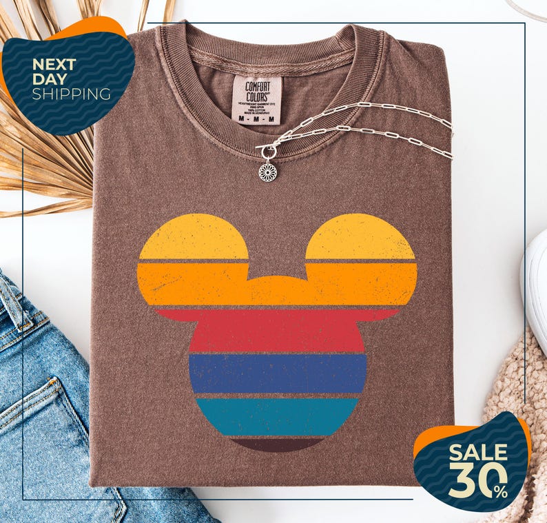 a brown t-shirt with a colorful Mickey Mouse design, along with a denim jacket and a necklace. The t-shirt is displayed on a white background, and there are two sale signs indicating a 30% discount.