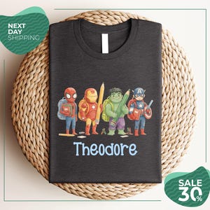 A black t-shirt with a graphic design featuring four cartoon characters, including Spider-Man, Iron Man, Captain America, and Theodore, along with the name Theodore printed on the shirt.