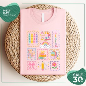 A pink t-shirt with a colorful graphic design featuring various school-related items such as crayons, a backpack, a pencil, and a rainbow. The t-shirt is displayed on a woven basket, and there are two green circular signs in the image.