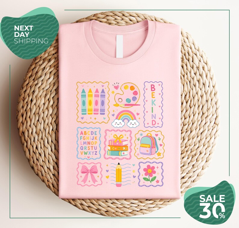 A pink t-shirt with a colorful graphic design featuring various school-related items such as crayons, a backpack, a pencil, and a rainbow. The t-shirt is displayed on a woven basket, and there are two green circular signs in the image.