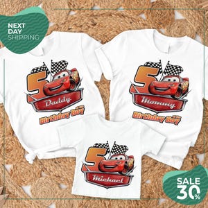 three white t-shirts with a Cars-themed design, including a red race car and a checkered flag. The t-shirts are labeled with the names "Daddy", "Michael", and "Mommy" and are displayed on a bed of straw.