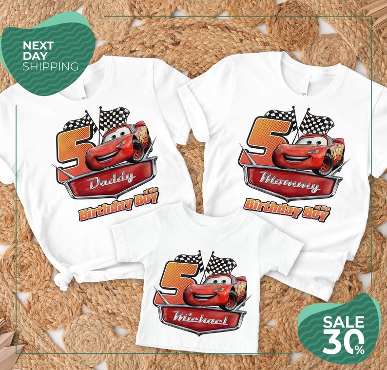 three white t-shirts with a Cars-themed design, including a red race car and a checkered flag. The t-shirts are labeled with the names "Daddy", "Michael", and "Mommy" and are displayed on a bed of straw.