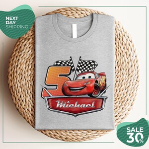 A gray t-shirt with a graphic design featuring a red car and checkered flags, along with the text "Michael" and the number "5". The t-shirt is displayed on a woven placemat, and there are two green circular sale signs in the image.