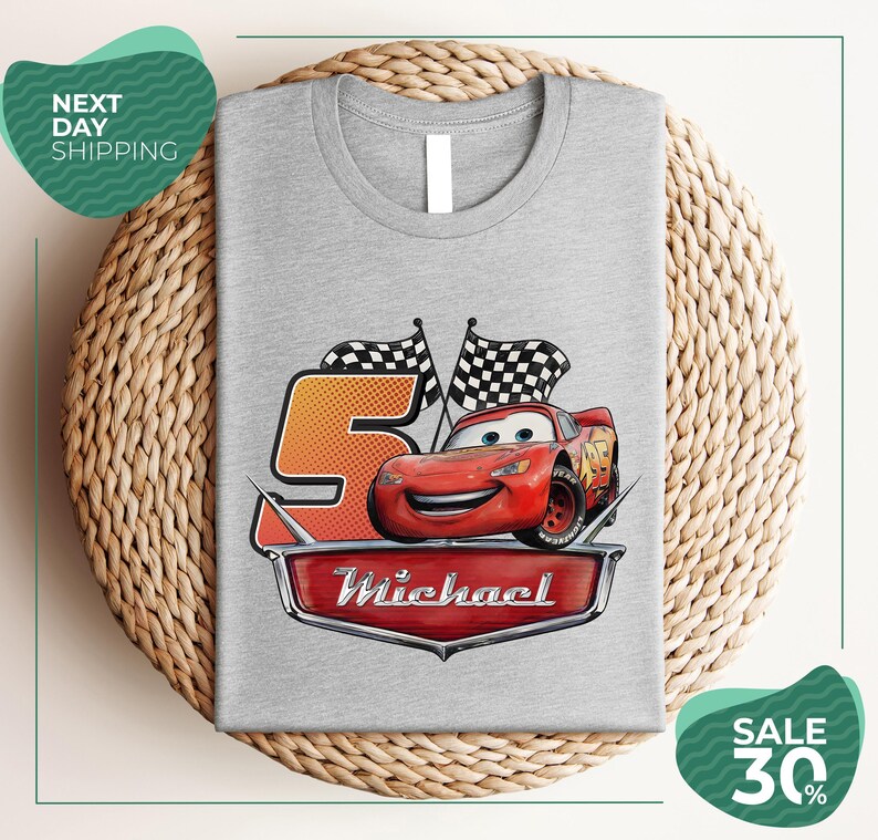 A gray t-shirt with a graphic design featuring a red car and checkered flags, along with the text "Michael" and the number "5". The t-shirt is displayed on a woven placemat, and there are two green circular sale signs in the image.