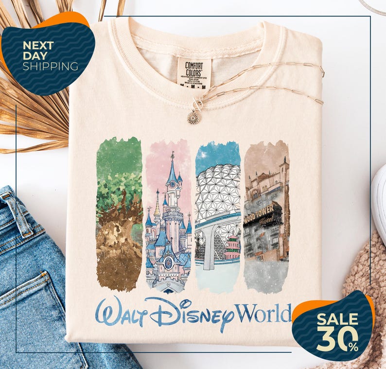 A white t-shirt with a graphic design featuring illustrations of various Disney landmarks, including the castle, the Eiffel Tower, and the Hollywood sign. The t-shirt is displayed on a white background, and there are jeans and a sweater visible in the background.