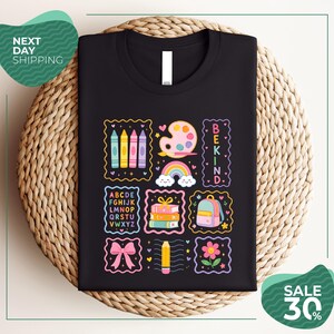 a black t-shirt with a colorful and whimsical design, including various school-related items such as pencils, books, and a backpack, along with the text "BEKIND" and "SALE 30%".