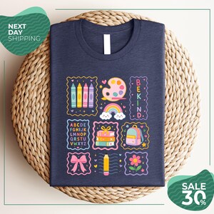 a dark blue t-shirt with a colorful and whimsical design, including various school-related items such as crayons, a backpack, a pencil, and a rainbow. The t-shirt is displayed on a woven basket, and there are two green circular signs in the image, one reading "Next Day Shipping" and the other indicating a sale with a 30% discount.