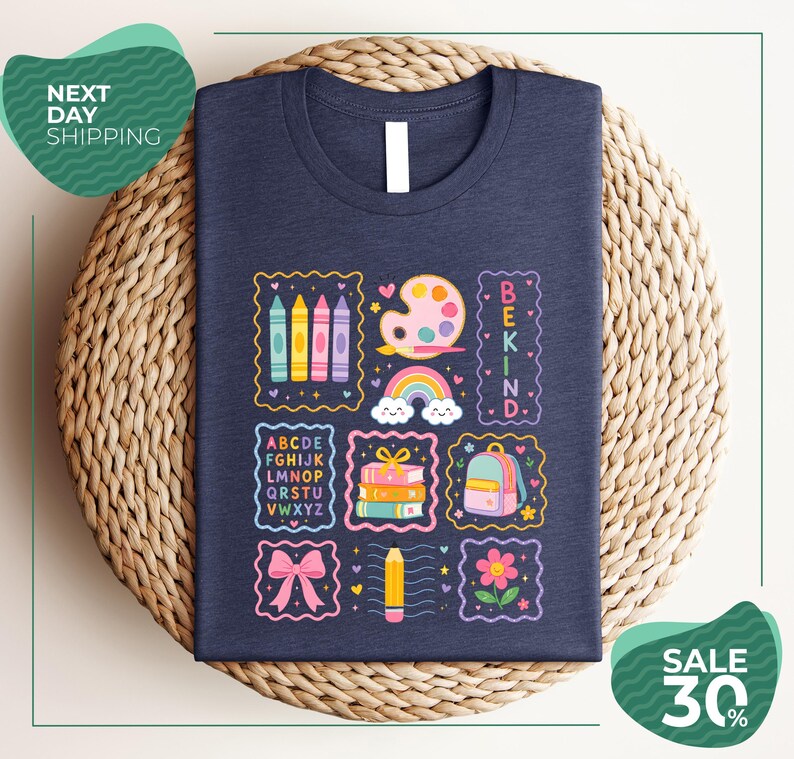 a dark blue t-shirt with a colorful and whimsical design, including various school-related items such as crayons, a backpack, a pencil, and a rainbow. The t-shirt is displayed on a woven basket, and there are two green circular signs in the image, one reading "Next Day Shipping" and the other indicating a sale with a 30% discount.
