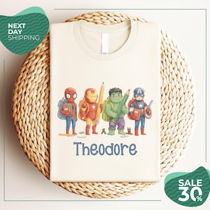 A white t-shirt with a graphic design featuring four cartoon characters, including Spider-Man, Iron Man, Hulk, and Captain America, along with the name Theodore. The t-shirt is displayed on a woven basket, and there are two green circular signs in the image, one indicating "Next Day Shipping" and the other promoting a "SALE 30%".
