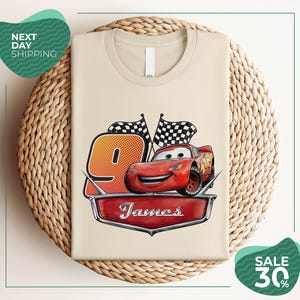 A t-shirt with a design featuring a red car and checkered flags, along with the text "James" and the number "9". The t-shirt is displayed on a woven basket, and there are two green circular signs in the background, one of which is promoting a sale of 30% off.