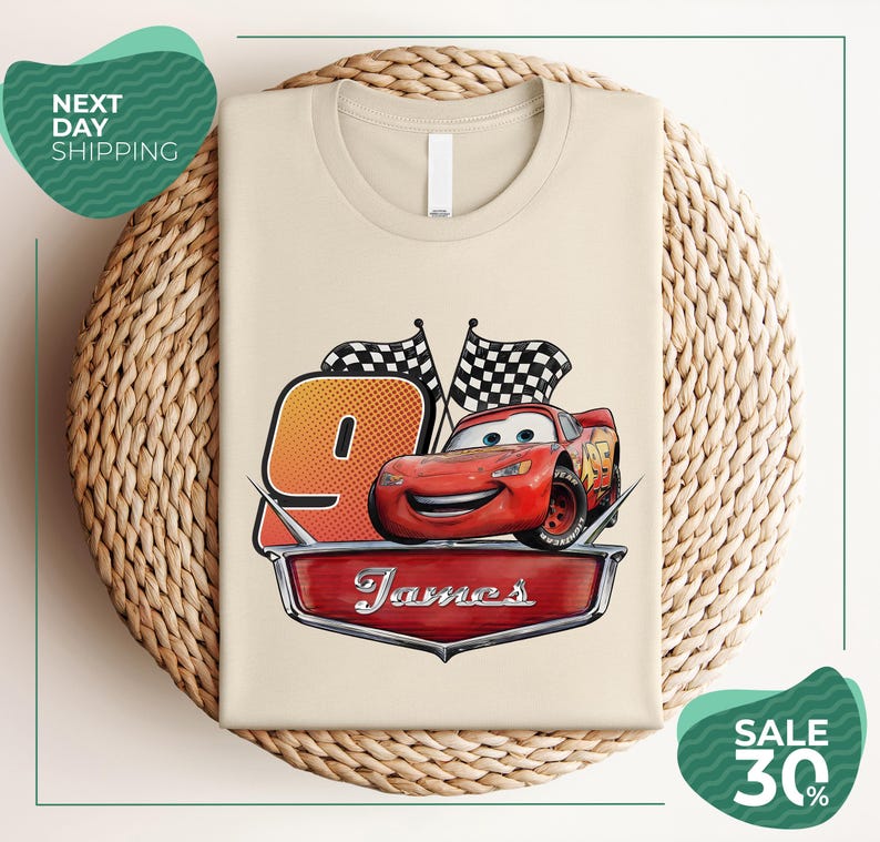 A t-shirt with a design featuring a red car and checkered flags, along with the text "James" and the number "9". The t-shirt is displayed on a woven basket, and there are two green circular signs in the background, one of which is promoting a sale of 30% off.