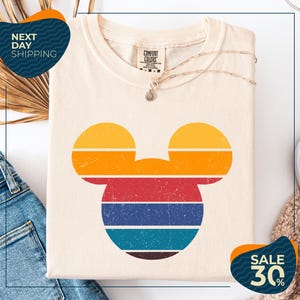 a white t-shirt with a colorful Mickey Mouse design, along with a pair of blue jeans and a beige sweater. The t-shirt is displayed on a white background, and there are two sale signs indicating a 30% discount.