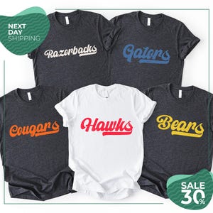 May include: A collection of five short-sleeved t-shirts. Four shirts are dark heather gray with the text Razorbacks, Gators, Cougars, and Bears printed in white, blue, orange, and yellow. One white shirt features the text Hawks in red.