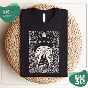 a black t-shirt with a white graphic design depicting a cat surrounded by various flora and fauna, including a bird and a butterfly. The t-shirt is displayed on a woven placemat, and there are two green circular sale signs in the image.