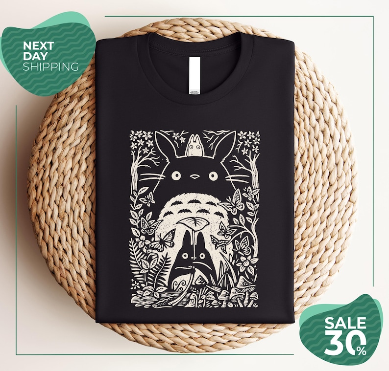 a black t-shirt with a white graphic design depicting a cat surrounded by various flora and fauna, including a bird and a butterfly. The t-shirt is displayed on a woven placemat, and there are two green circular sale signs in the image.