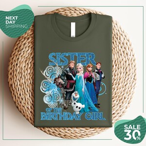 A green t-shirt with a graphic design featuring characters from the Disney movie Frozen, specifically the sisters Elsa, Anna, Olaf, and Olaf.