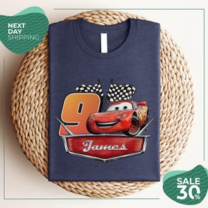 A blue t-shirt with a graphic design featuring a red car and checkered flags, along with the text "James" and the number "9". The t-shirt is displayed on a woven placemat, and there are two green sale signs in the image.