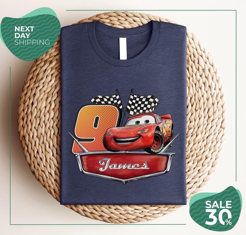 A blue t-shirt with a graphic design featuring a red car and checkered flags, along with the text "James" and the number "9". The t-shirt is displayed on a woven placemat, and there are two green sale signs in the image.