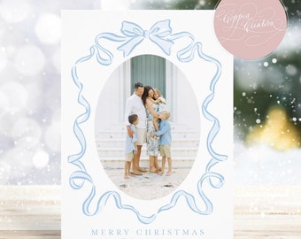 May include: A white Christmas card with a family photo inside an oval frame, adorned with light blue ribbon and bow details. The text "MERRY CHRISTMAS" and family name are printed at the bottom. The card is set on a wooden surface.