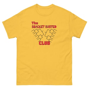 May include: Mustard yellow t-shirt with the text "The Bracket Busted Club" in red and black. The graphic features a bracket design. The shirt is made of a soft material.