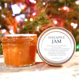 May include: A jar of pineapple jam with the lid off, next to the jar's lid. The label reads "Pineapple Jam" and lists ingredients. The jam is a light orange color. The jar is 6 oz.