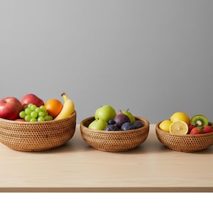 Op de afbeelding: Drie geweven rotan fruitschalen gevuld met vers fruit. De schalen bevatten appels, druiven, sinaasappels, bananen, pruimen, citroenen, kiwi's en aardbeien. De schalen zijn gerangschikt op een licht houten oppervlak.