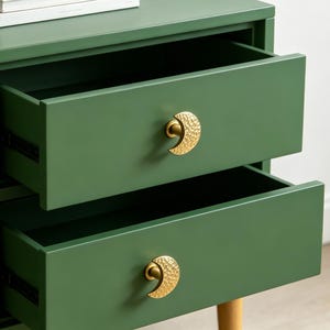 May include: A dark green wooden chest of drawers featuring two open drawers with gold-toned, crescent moon-shaped knobs that have a hammered texture. A stack of two white books rests on the flat top surface of the furniture piece.