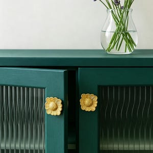 Puede incluir: Un armario de color verde oscuro con dos puertas que presentan paneles de vidrio estriado vertical y pomos dorados en forma de flor. Un jarrón de vidrio transparente con tallos de lavanda morada descansa sobre la superficie superior.