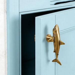 Peut inclure: Un bouton de placard en métal couleur laiton en forme de requin, fixé sur une porte de placard en bois peinte en bleu clair. La figurine de requin est orientée verticalement, tête vers le haut, avec des détails de nageoires et de branchies.