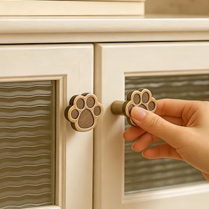 May include: A cabinet knob shaped like a paw print with a dark brown wood inlay and a metallic gold-toned border. The knob is mounted on a light-colored wooden cabinet door featuring a glass panel with a horizontal wavy texture pattern.