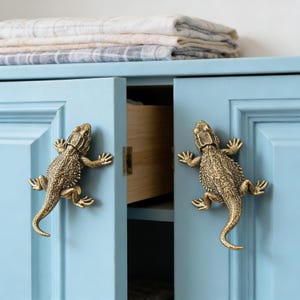 May include: A light blue cabinet featuring two brass-coloured lizard-shaped drawer pulls. The cabinet has a partially open drawer revealing a light wood interior, and a stack of folded, neutral-toned plaid textiles rests on the top surface.