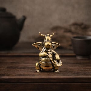 May include: A small, metallic brass-colored figurine of a dragon with wings, horns, and a rounded belly. The dragon is depicted in a standing position, holding a long, thin object in its hands, featuring a textured surface and a warm, golden finish.